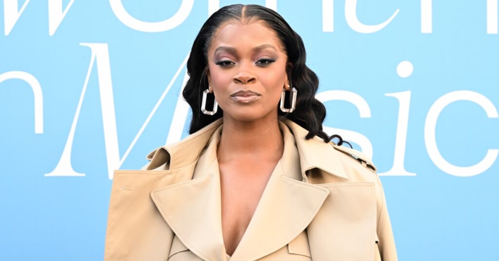 Ari Lennox poses in a beige trench coat with large hoop earrings against a blue background.