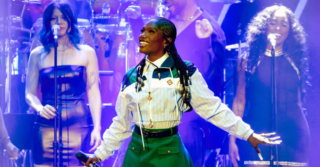 Doechii in a green skirt and white shirt performs on stage with two backup singers in black dresses, under blue lighting.
