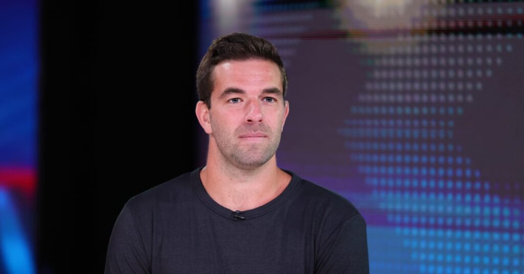 Fyre Festival founder Billy McFarland in a black t-shirt is standing in front of a digital screen with a dotted pattern.