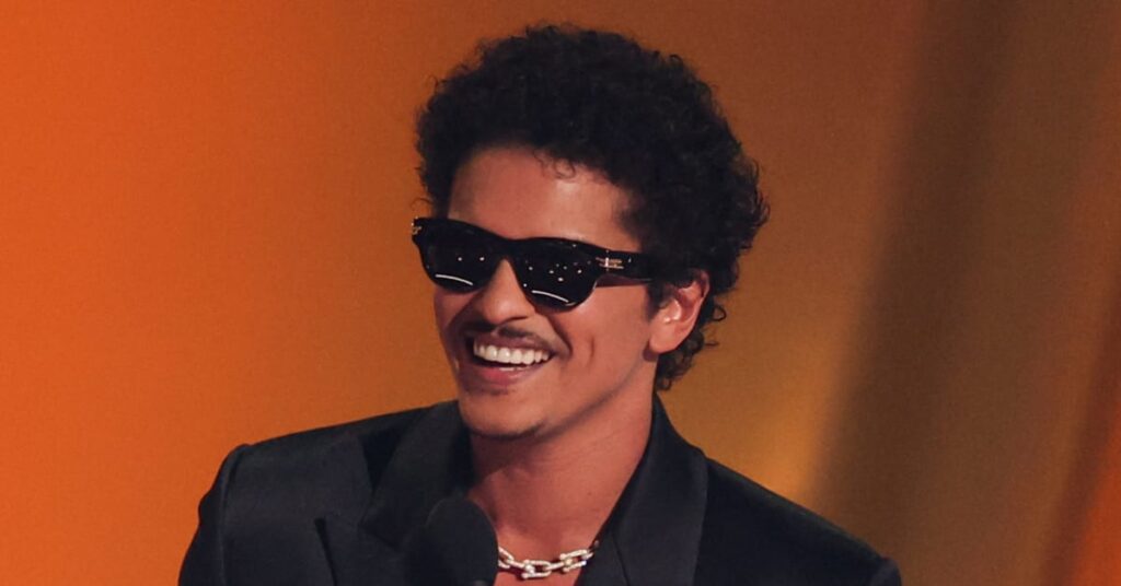 Bruno Mars, wearing sunglasses and a dark suit, smiles at an event.
