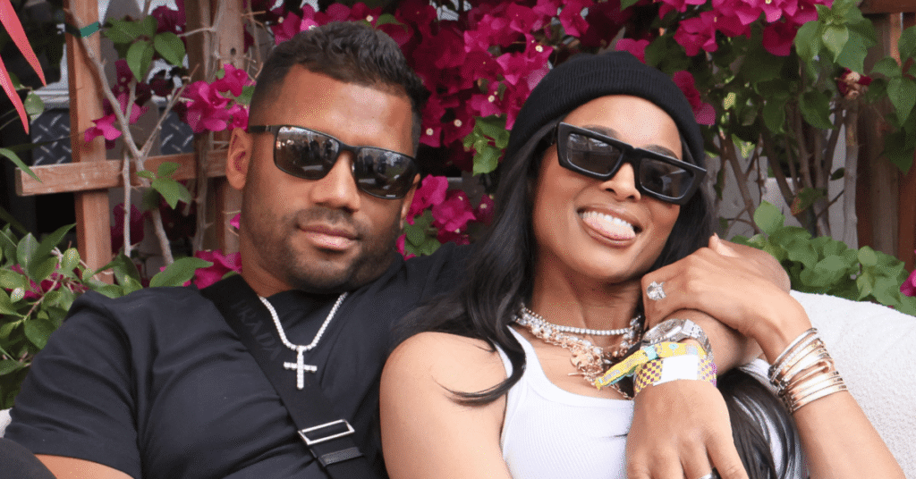 Russell Wilson and Ciara are sitting together, both wearing sunglasses. Ciara is smiling and leaning on Russell, with flowers in the background.