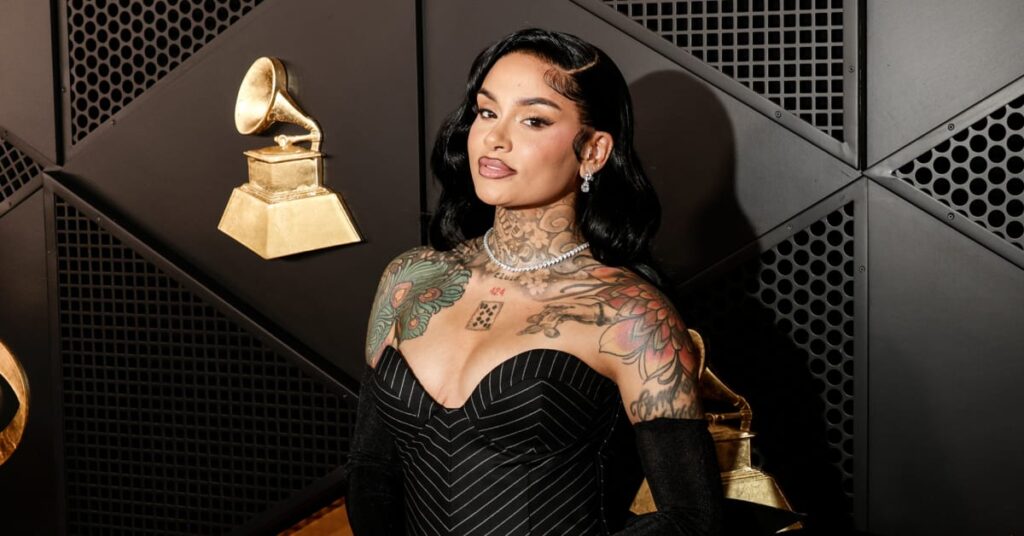 Kehlani at an event, wearing a pinstripe dress. She has visible tattoos and is posing in front of Grammy-themed decor.