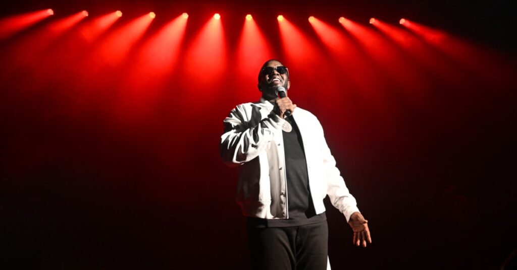 Diddy wearing sunglasses and a white jacket performs on stage under red lights.