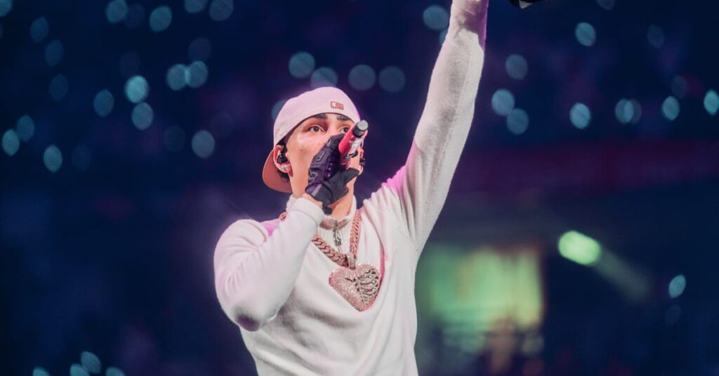 Omar Courtz on stage wearing a white cap, gloves, and a heart-shaped necklace, holding a microphone, with an arm raised in a lively atmosphere.