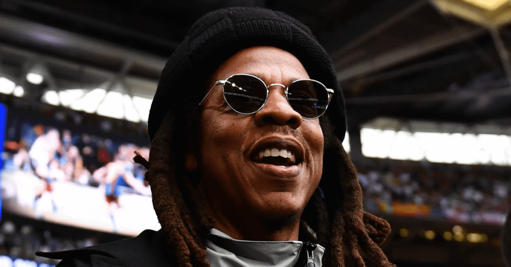 A person wearing sunglasses and a beanie, smiling in a stadium setting.
