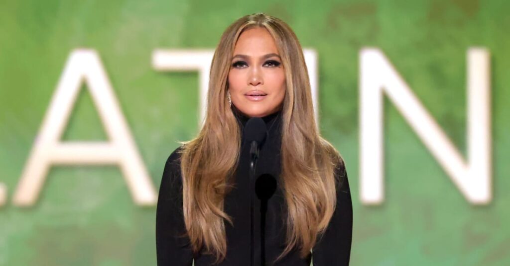 Jennifer Lopez speaking at an event, wearing a black outfit, with long wavy hair, against a green background.