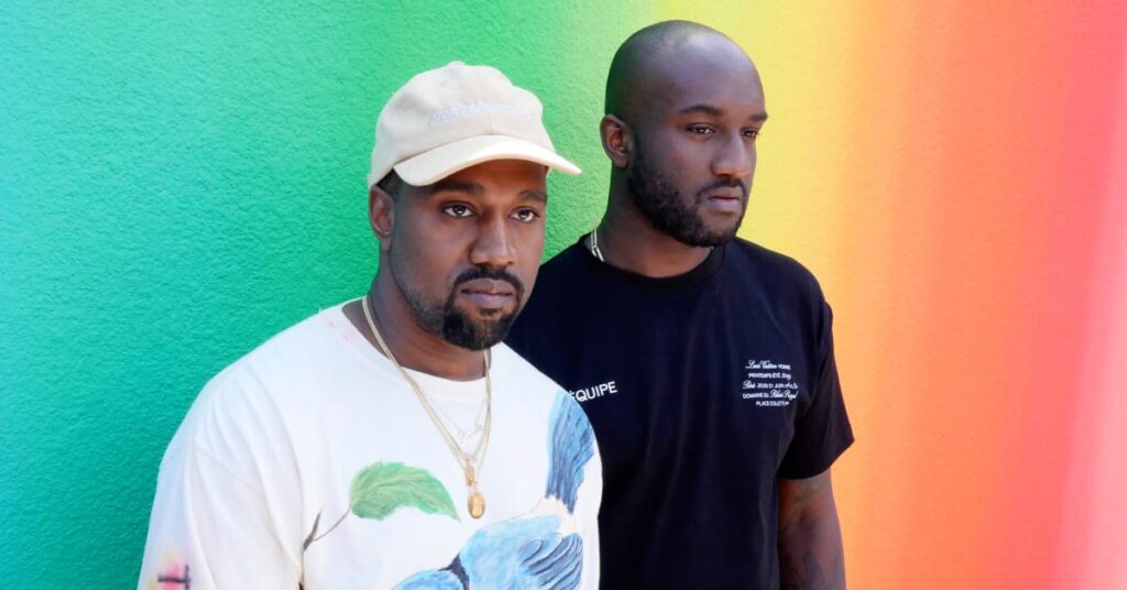 Kanye West and Virgil Abloh stand in front of a colorful gradient wall. Kanye wears a bird-print shirt and cap, Virgil in black tee.