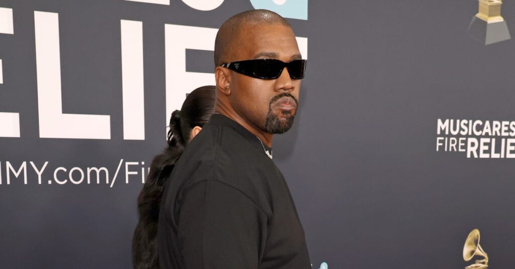Kanye West wearing sunglasses and a black outfit at an event, standing in front of a backdrop with text and logos.