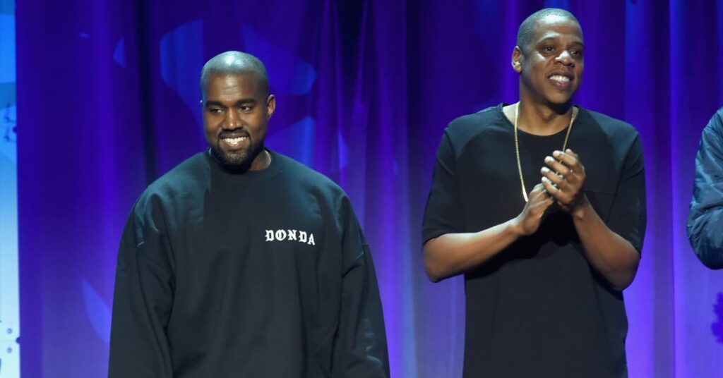 Kanye West and Jay-Z stand on stage, smiling. Kanye wears a "Donda" sweatshirt, and Jay-Z claps, wearing a black shirt.
