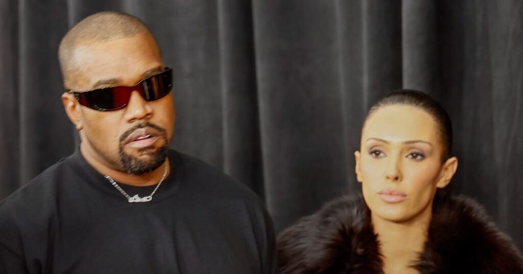 Ye in sunglasses and a black shirt stands next to wife Bianca Censori with slicked-back hair wearing a dark fur coat, against a dark curtain backdrop.