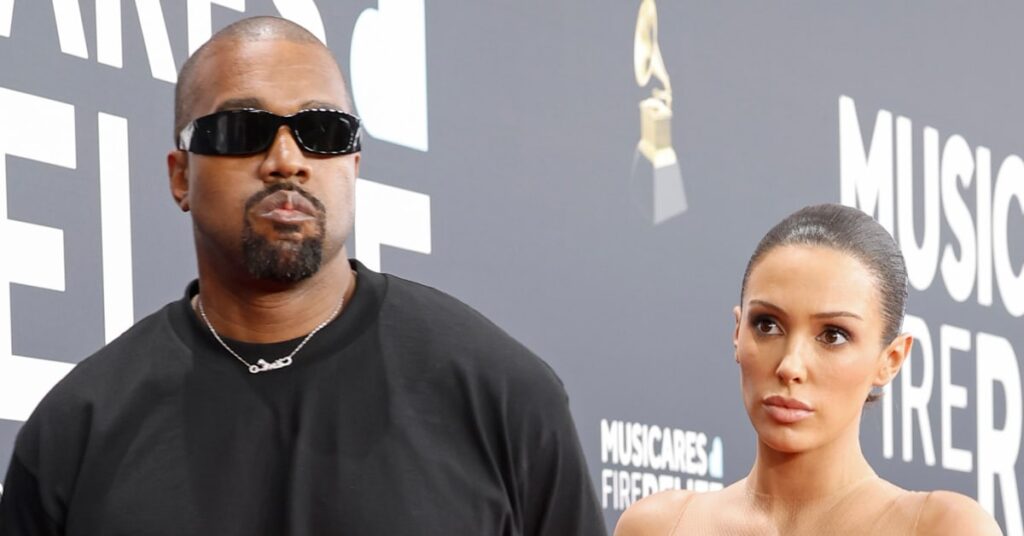 Kanye West wearing sunglasses and a black shirt stands beside Bianca Censori in a beige outfit at an event with a black backdrop.