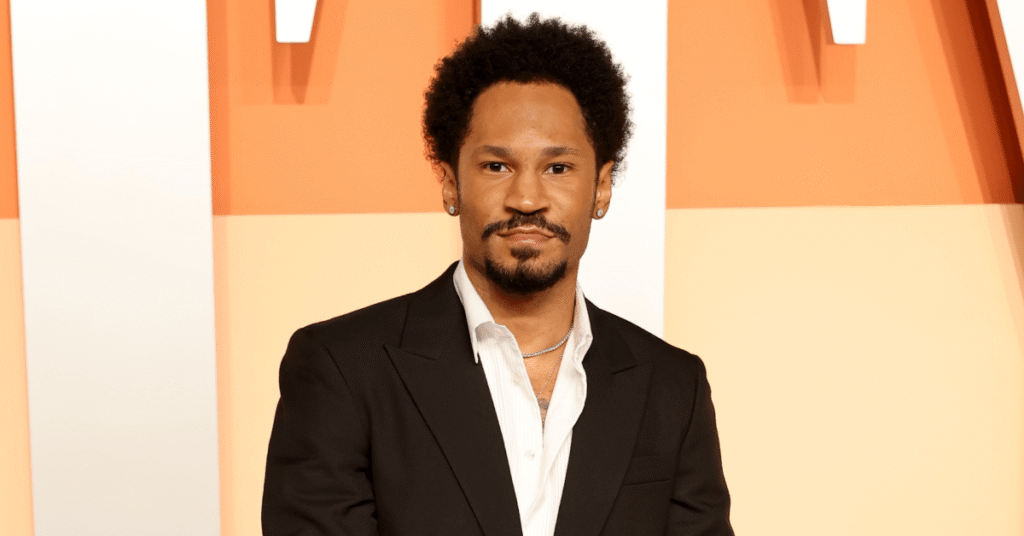 A man with short curly hair and a beard, wearing a black suit and white shirt, stands against an orange and white background.