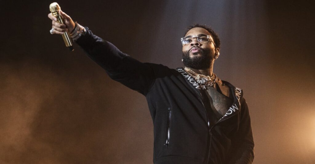 Kevin Gates on stage holding a microphone, wearing glasses and a black jacket, with dramatic lighting in the background.