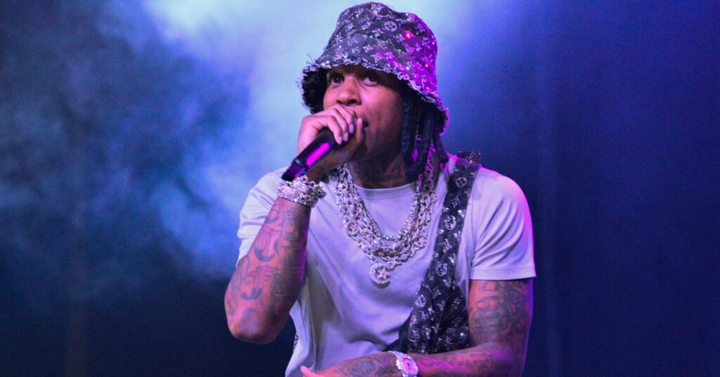 Lil Durk performing on stage, wearing a patterned bucket hat and white shirt, with tattoos and multiple chains.