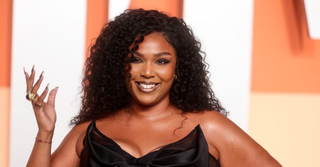 Lizzo poses in a black dress with a bow detail, smiling with her hand raised, against an orange and white background.
