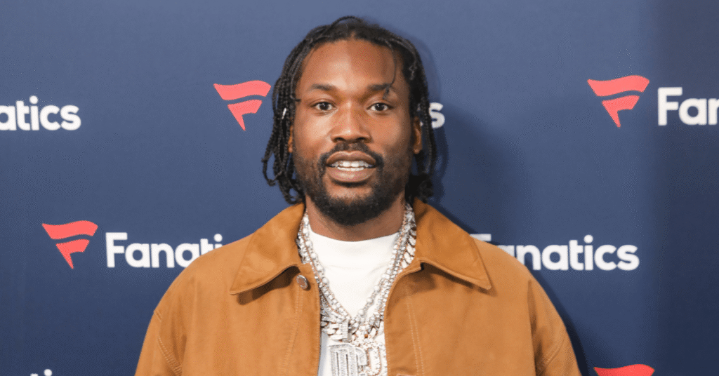 Meek Mill at a Fanatics event, wearing a brown jacket and a large chain necklace, smiling at the camera.