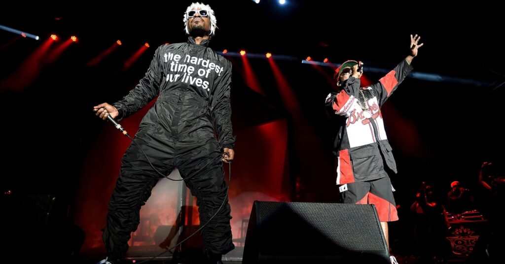 André 3000 and Big Boi performing on stage as OutKast, wearing distinctive outfits under bright stage lights.