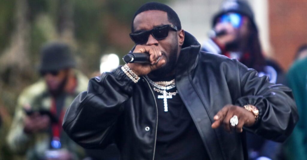 Diddy wearing a black leather jacket and sunglasses performs on stage, holding a microphone.