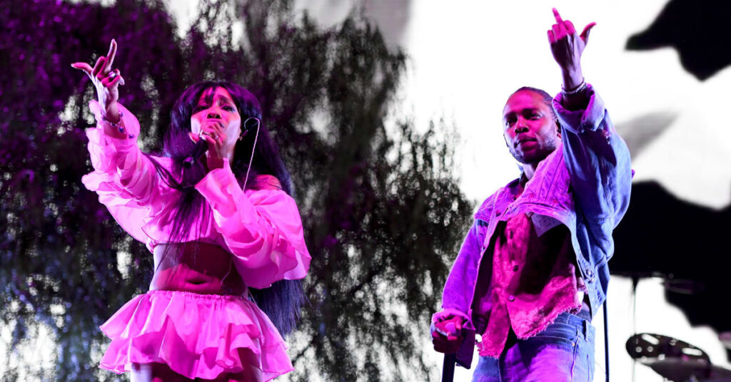 SZA and Kendrick Lamar performing on stage, both holding microphones and raising their middle fingers, with a dramatic backdrop.