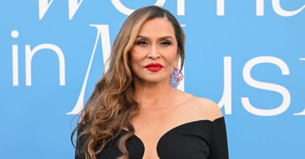 Tina Knowles with long wavy hair and red lipstick stands in front of a blue background with text. She's wearing a black dress.