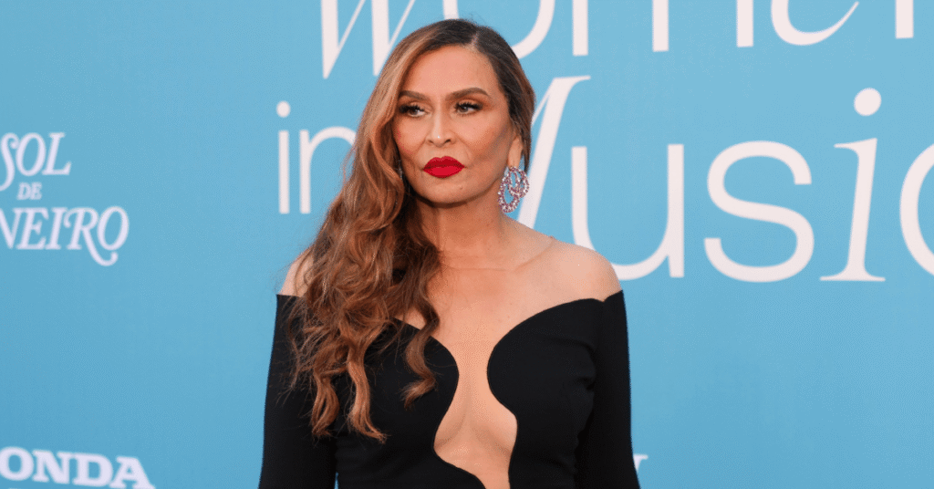 A woman with long hair in a black dress stands against a blue background with text, wearing red lipstick and earrings.
