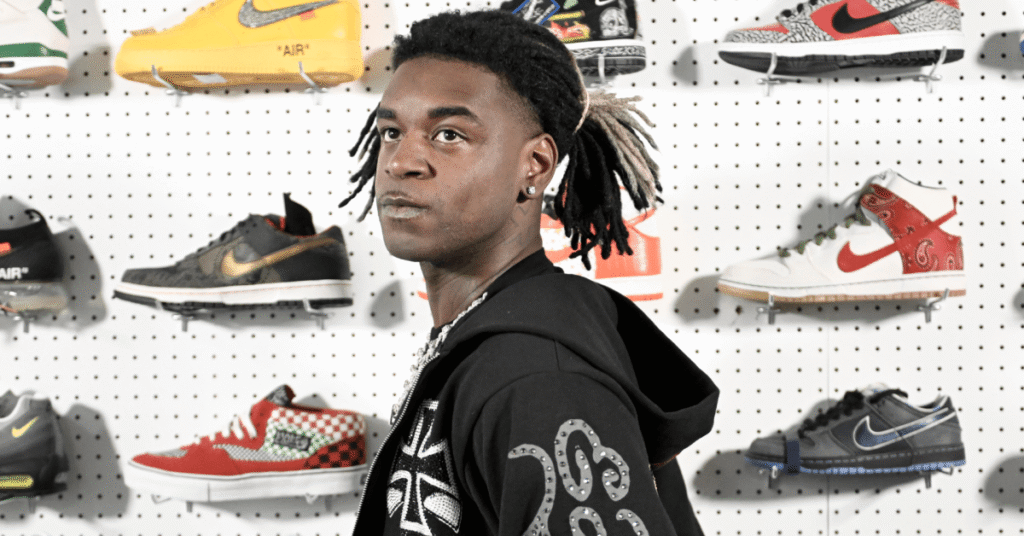 A man with dreadlocks stands in front of a wall displaying various colorful sneakers. He wears a black hoodie with white designs.