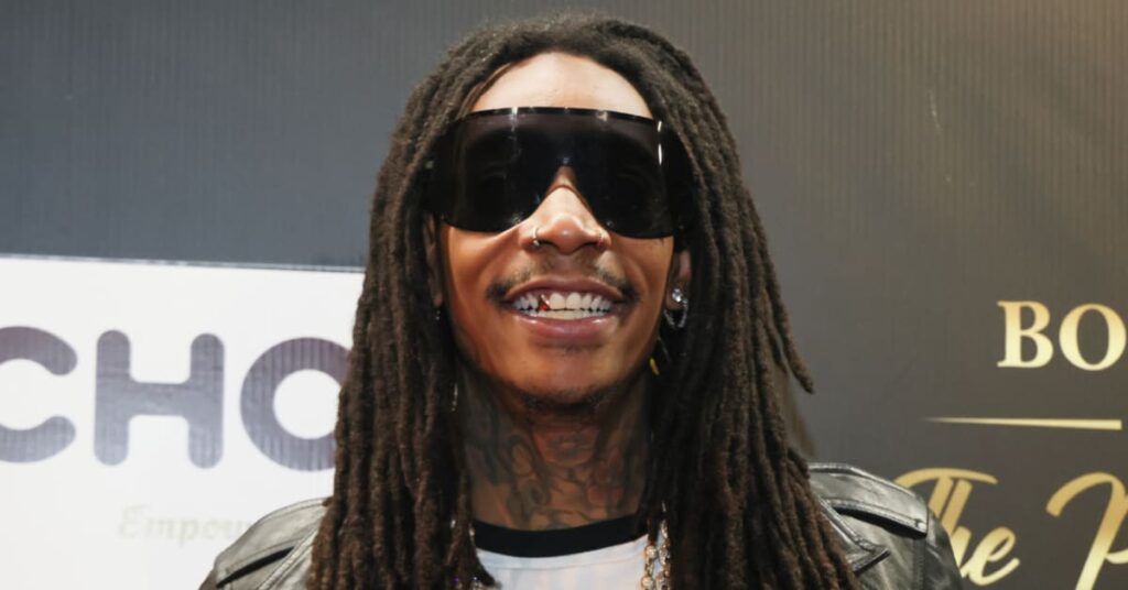 Rapper Wiz Khalifa wearing sunglasses and a leather jacket, smiling at an event.