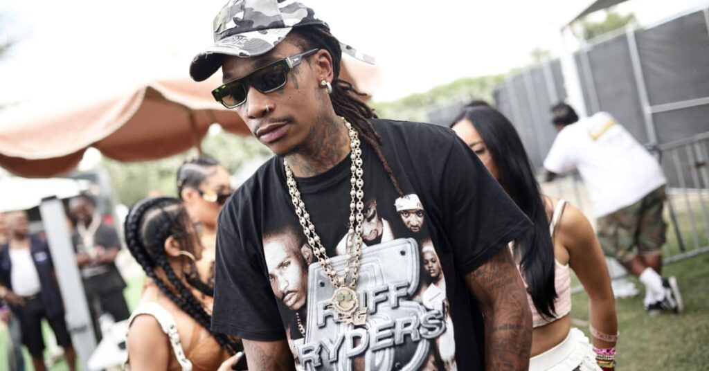 Wiz Khalifa wearing sunglasses, a camo cap, and a Ruff Ryders T-shirt, with chains around his neck, stands outdoors at an event.