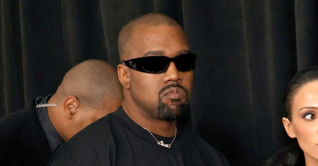 Kanye West wearing sunglasses and a black shirt, standing in front of a dark curtain.