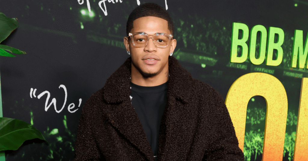 A man in a brown coat and glasses stands in front of a "Bob Marley One Love" backdrop.