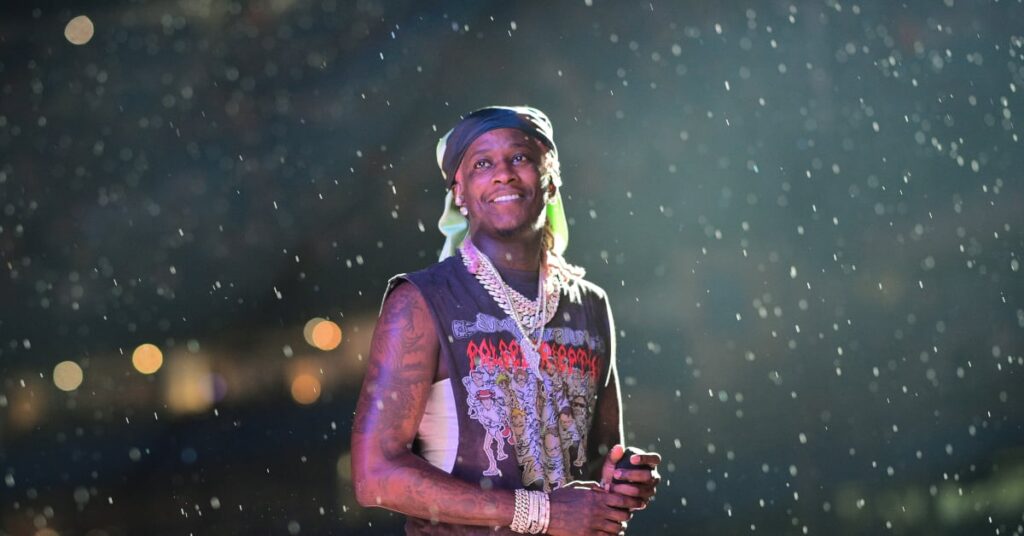 Young Thug on stage under rain, wearing a bandana and jewelry, holding a microphone and smiling, with blurred lights in the background.