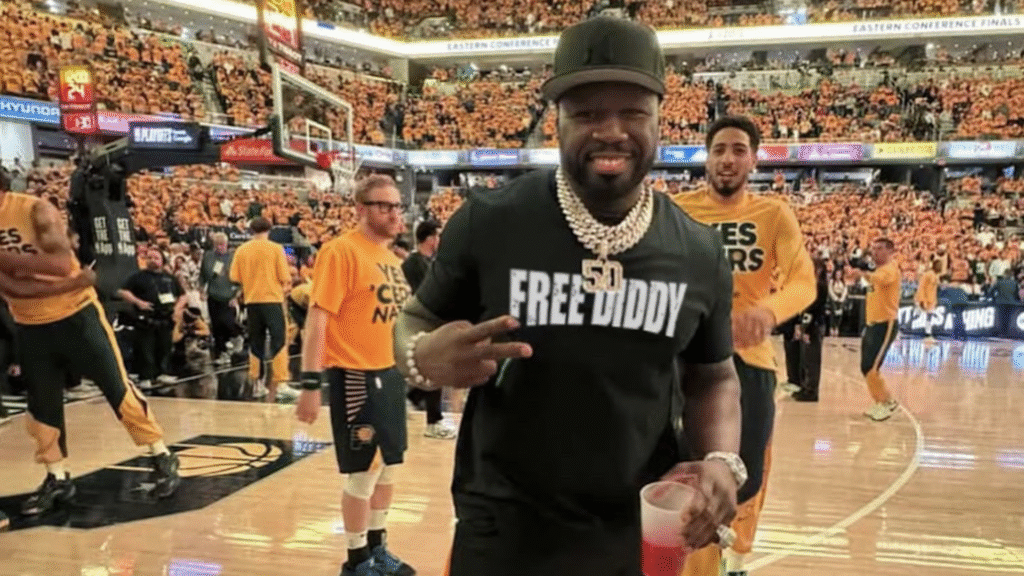50 Cent wearing a black cap and shirt, sitting courtside at a basketball game, surrounded by fans in yellow shirts.