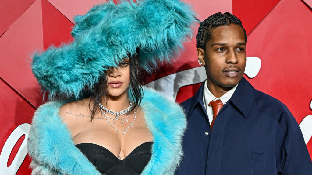 Rihanna in a blue fur outfit and hat, with ASAP Rocky in a navy coat and tie, posing at an event with a red backdrop.