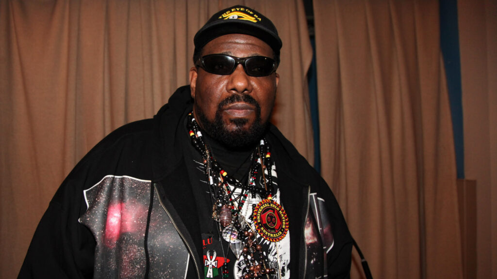 A man wearing sunglasses, a cap, and a jacket adorned with necklaces stands in front of a curtain.