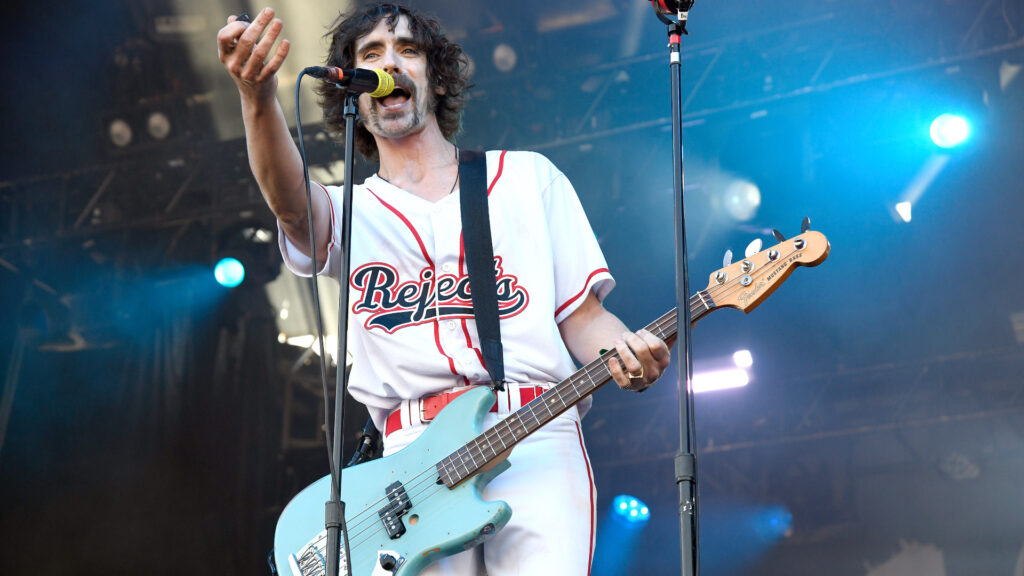Tyson Ritter on stage playing a light blue bass guitar, wearing a white "Rejects" baseball jersey, singing into a microphone.