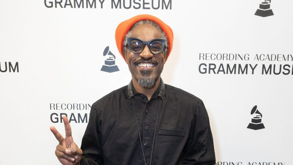 André 3000 at the Grammy Museum, wearing an orange hat, glasses, and a black shirt, smiling and holding up a peace sign.