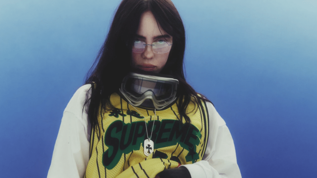 Billie Eilish wearing a Supreme jersey and goggles, with long dark hair and a serious expression against a blue background.