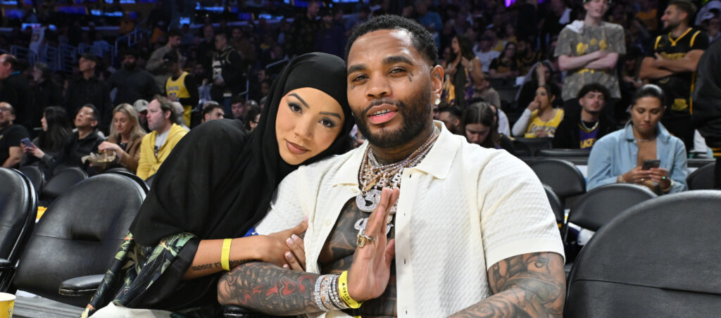 A man with tattoos and a woman in a hijab sit together courtside at a basketball game, surrounded by other spectators.