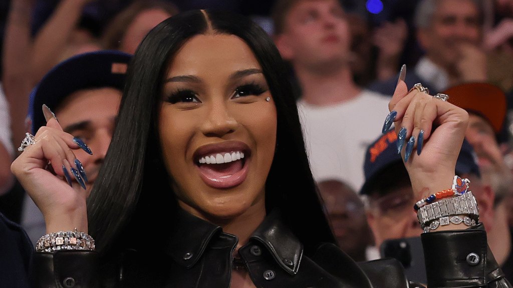 Cardi B smiling, wearing a black leather jacket and jewelry, with long nails. She is in a crowd, appearing excited.