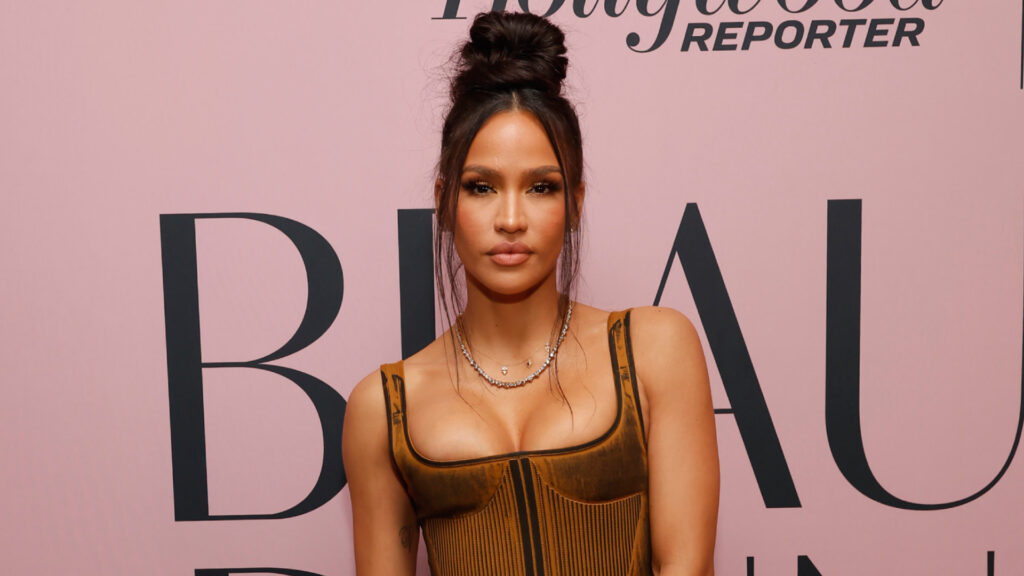 Cassie Reportedly Welcomes Third Child With Alex Fine Cassie Ventura circa 2003 with long hair in a high bun, wearing a brown corset top, stands in front of a pink backdrop with text.