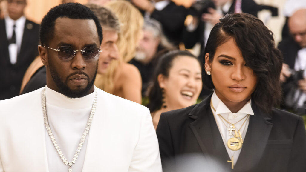 Sean "Diddy" Combs in a white suit and sunglasses, with Cassie Ventura in a black tuxedo, both at a formal event.
