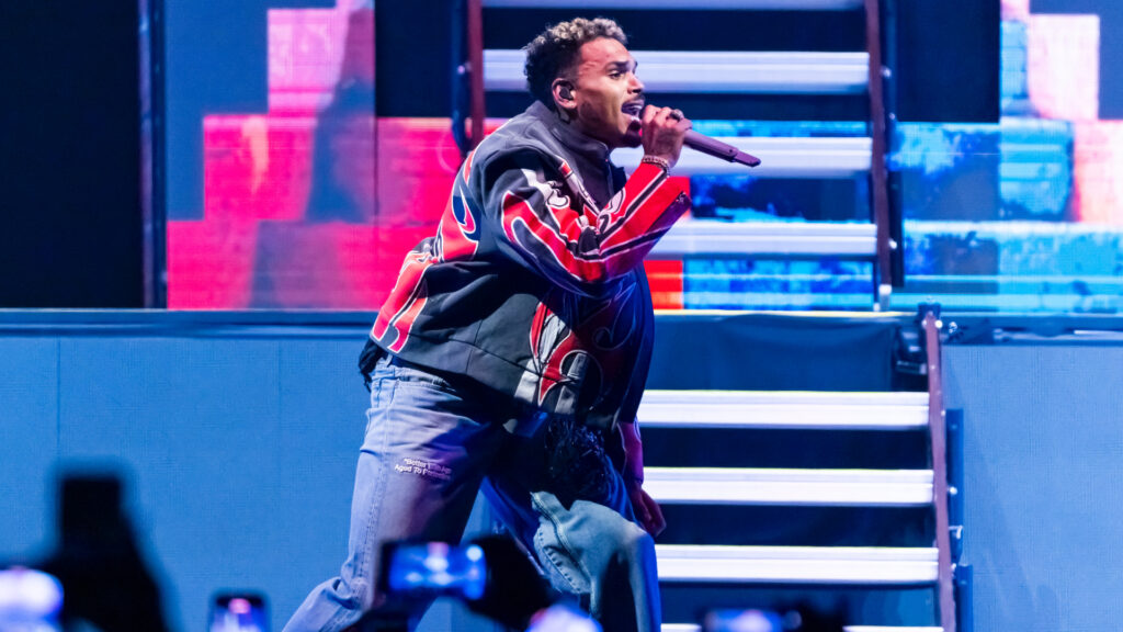 Chris Brown Reportedly Arrested for Allegedly Smashing Bo… Chris Brown in a colorful jacket performs energetically on stage, holding a microphone, with vibrant lighting and steps in the background.
