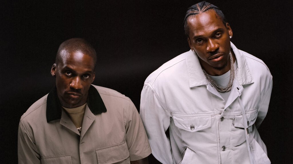 Clipse in stylish outfits pose against a dark background. Malice wears a beige jacket, Pusha T a white jacket with chains.