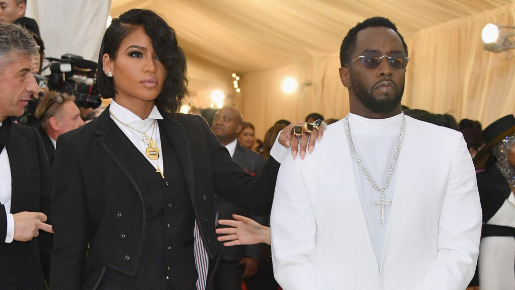 Diddy’s Team Plays Audio of Cassie Threatening to Kill Ma… Cassie in a black suit with gold jewelry and Sean "Diddy" Combs in a white suit with sunglasses at a formal event.