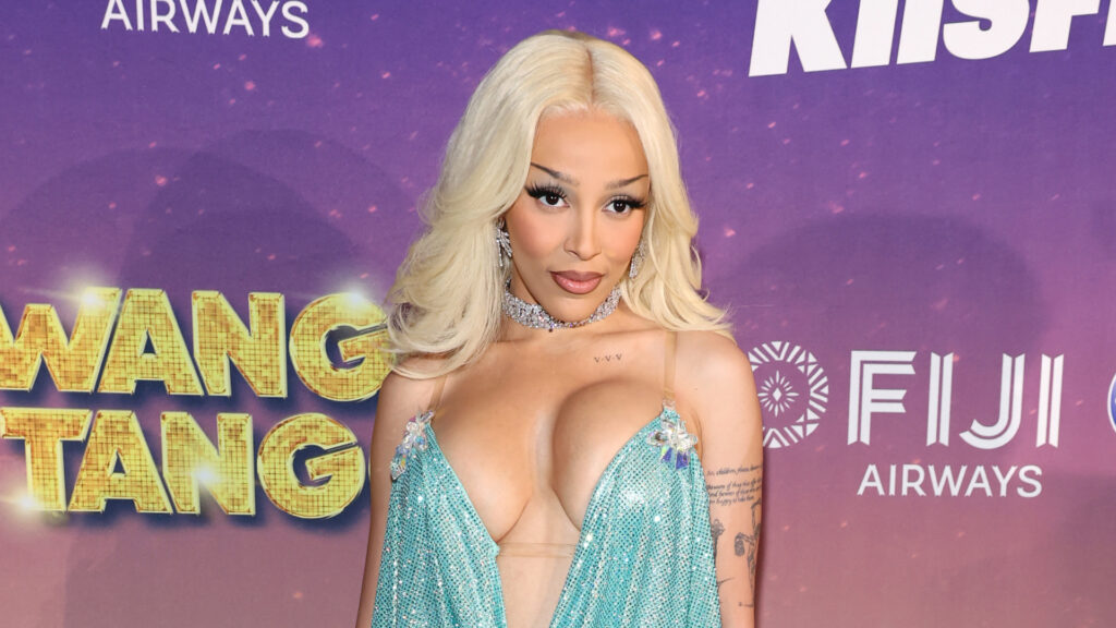 Doja Cat poses on the red carpet in a shimmering blue gown with a deep neckline, against a purple backdrop with logos.