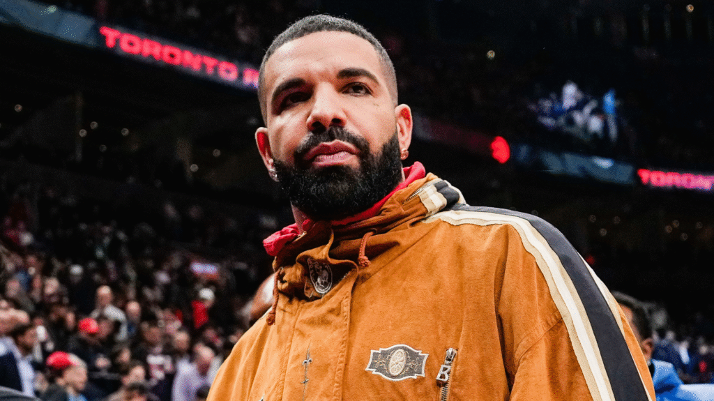 Drake at a sports event, wearing a brown jacket with a crowd in the background.