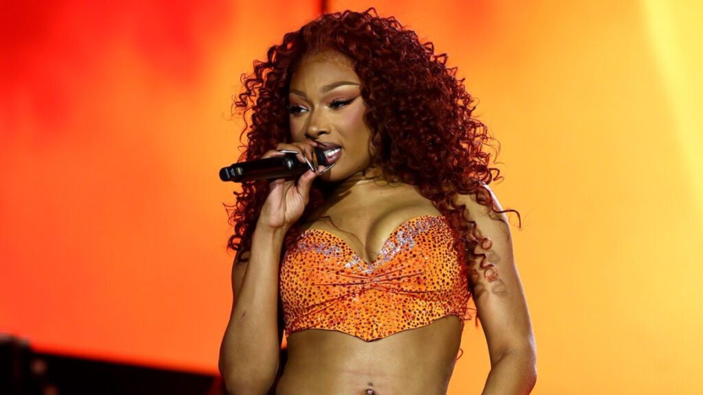 Megan Thee Stallion with curly red hair performs on stage, wearing an orange sparkly top, holding a microphone against a vibrant orange background.