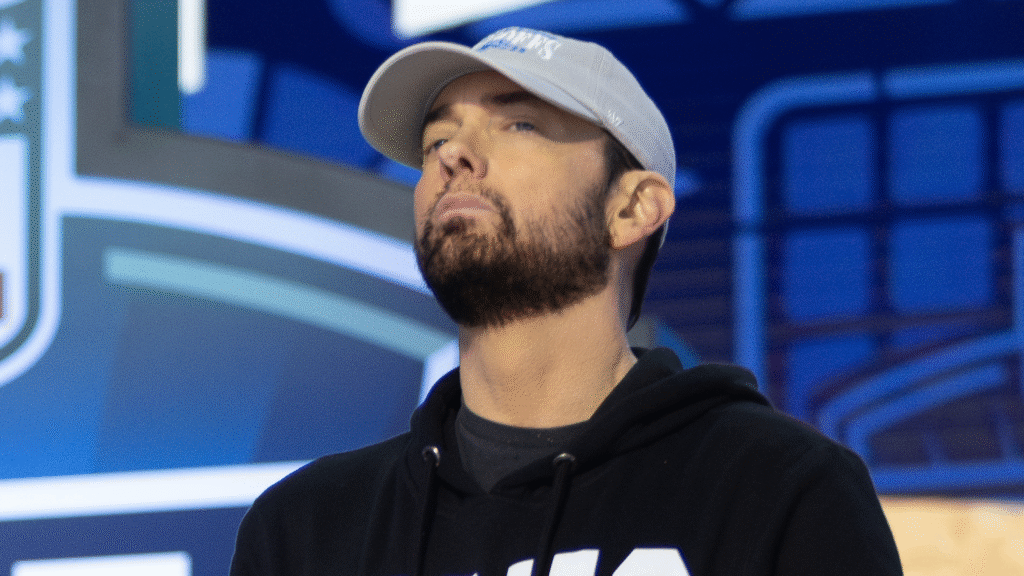 Eminem’s Publisher Sues Meta Over Unlicensed Use of Music… A man with a beard wearing a gray cap and black hoodie stands in front of a blurred blue background.