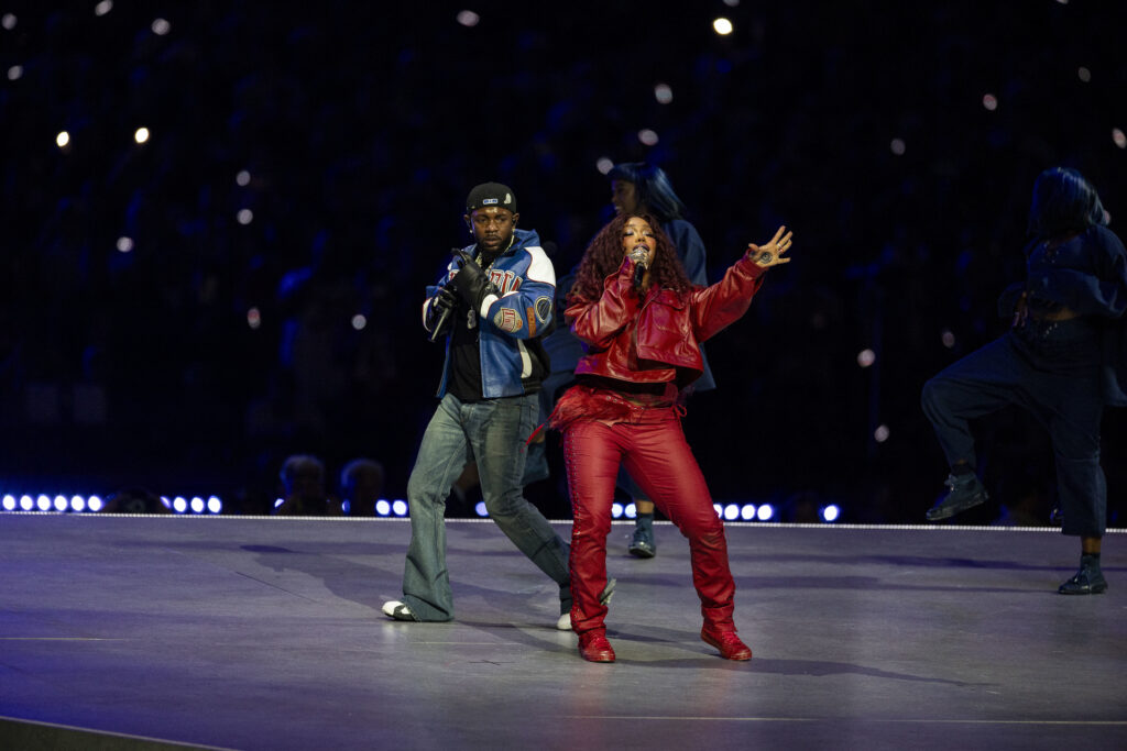 Every No. 1 Rap Song in Hot 100 History, Feat. Drake, Ken… SZA in a red outfit sings energetically on stage, accompanied by Kendrick Lamar in a blue jacket and jeans, with a dark background.