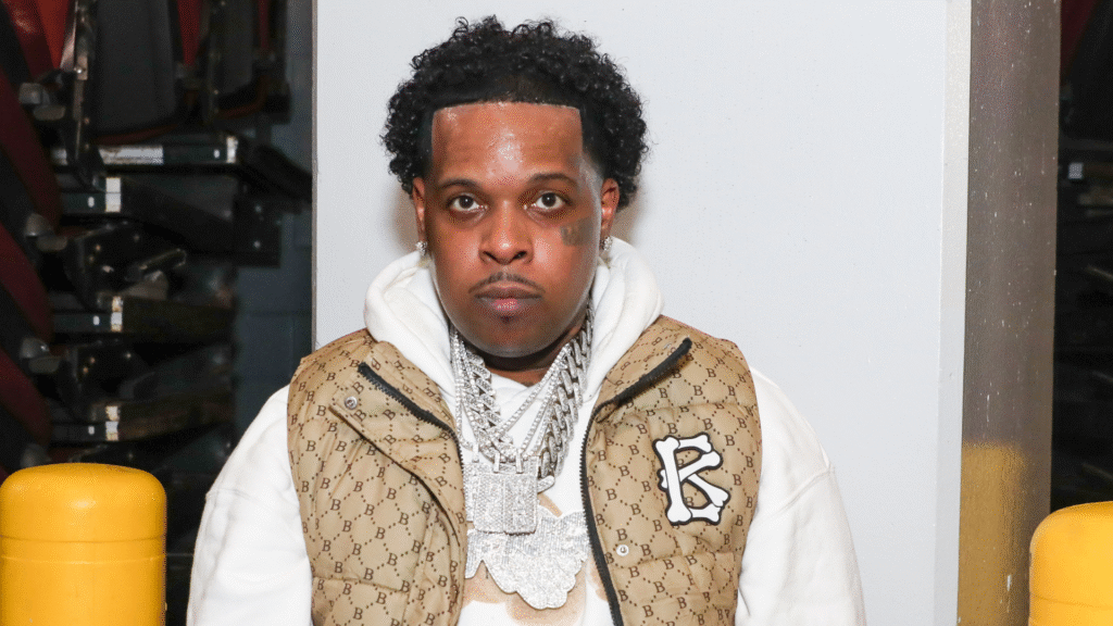 A man with short curly hair, wearing a brown patterned vest, white hoodie, and multiple silver chains, stands indoors.